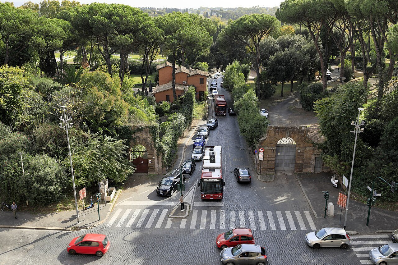 Traffic seen from above on a road running between trees past an old arched gateway