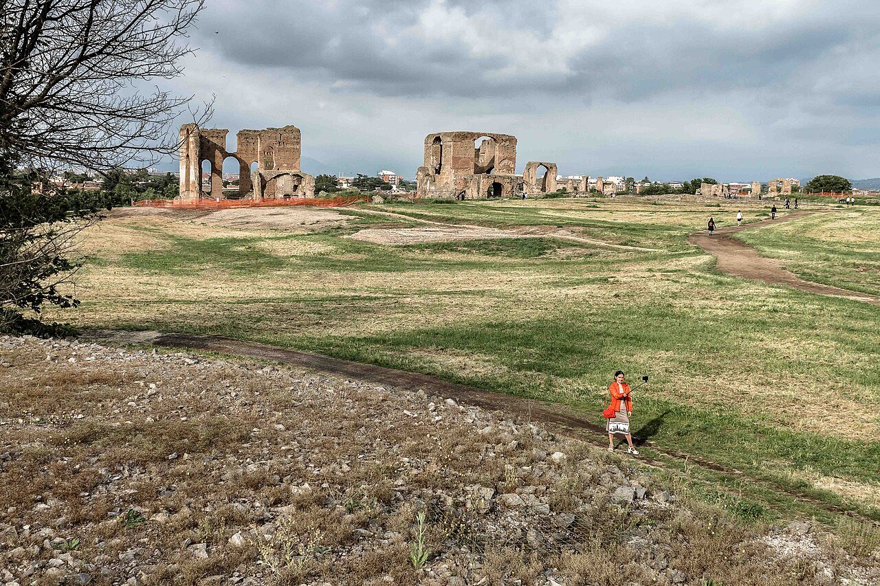 The ruins of the Villa of the Quintilii across a wide field, a visitor walking in the foreground