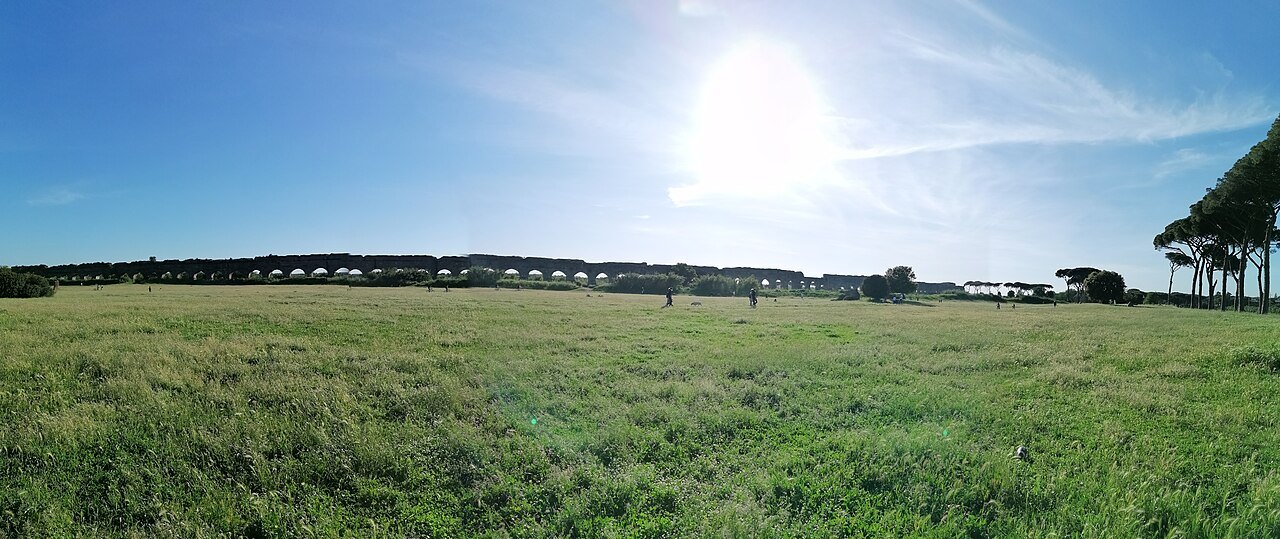 A wide green meadow under a bright sun, a line of aqueduct arches low on the horizon