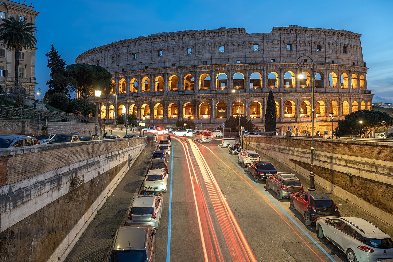 The Colosseum lit at dusk at the end of a road, car lights streaking below