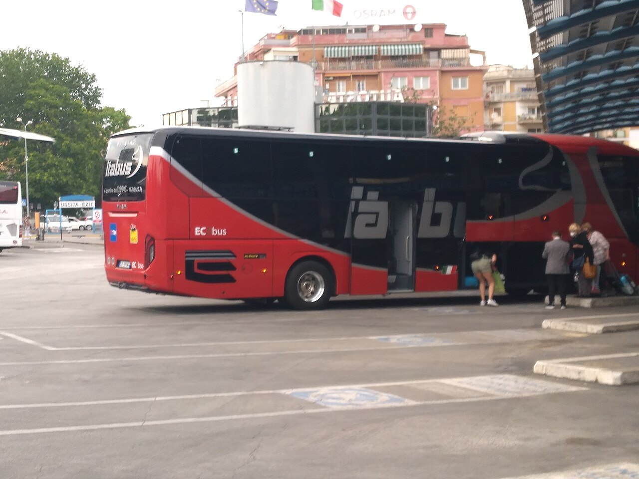A red long-distance coach at a bus terminal, passengers loading luggage