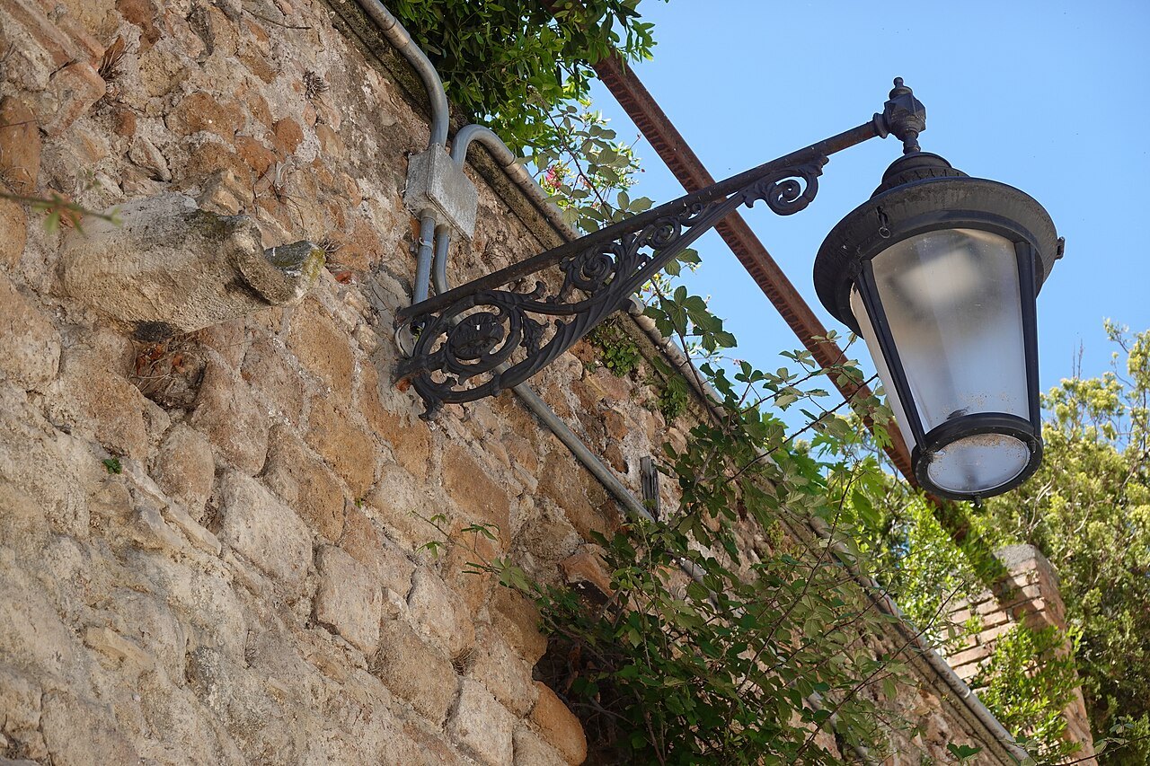 A wrought-iron lamp on a stone wall by the Tiber, leaves against the sky