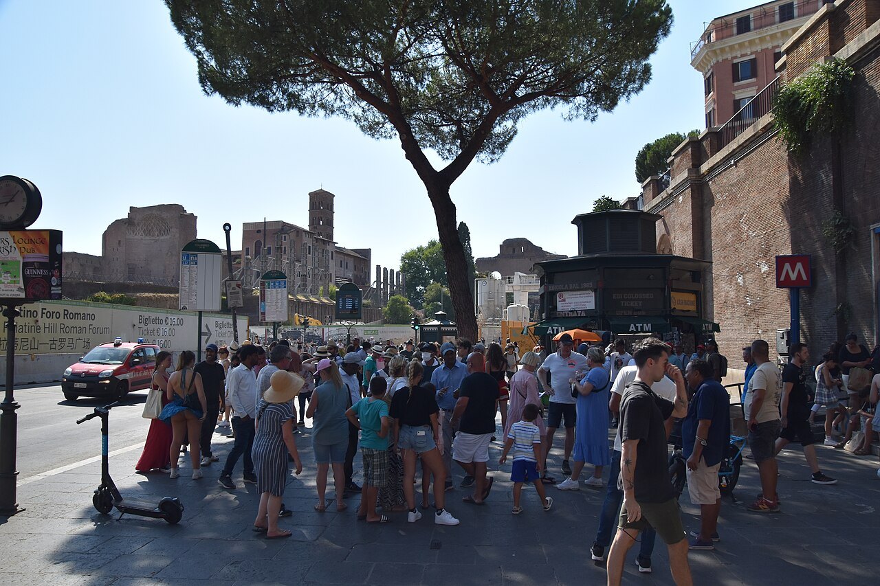 A crowd at the entrance to Colosseo station on Metro line B, under a pine and the ancient walls