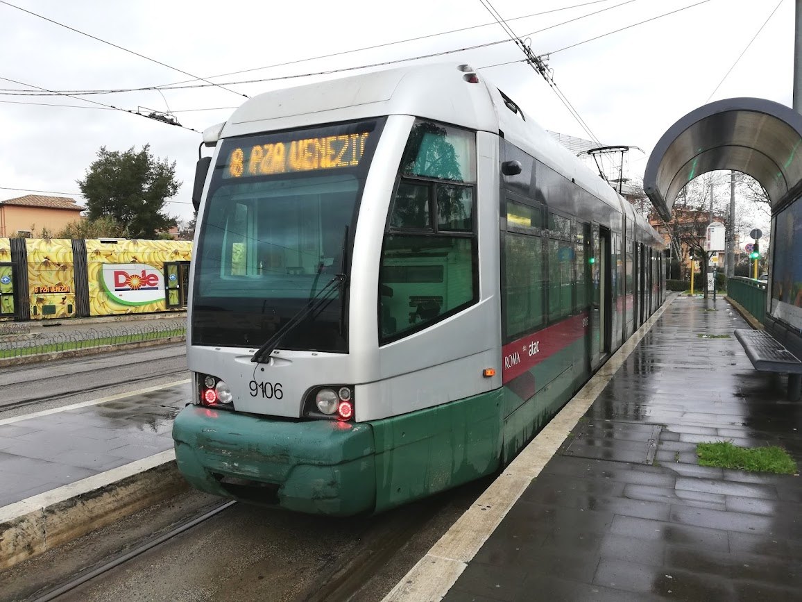 A modern tram at a stop on a wet day, its route shown on the front