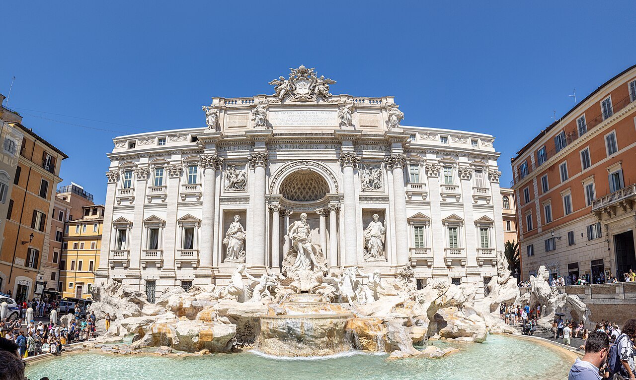 The Trevi Fountain and its crowds under a blue sky, houses on either side